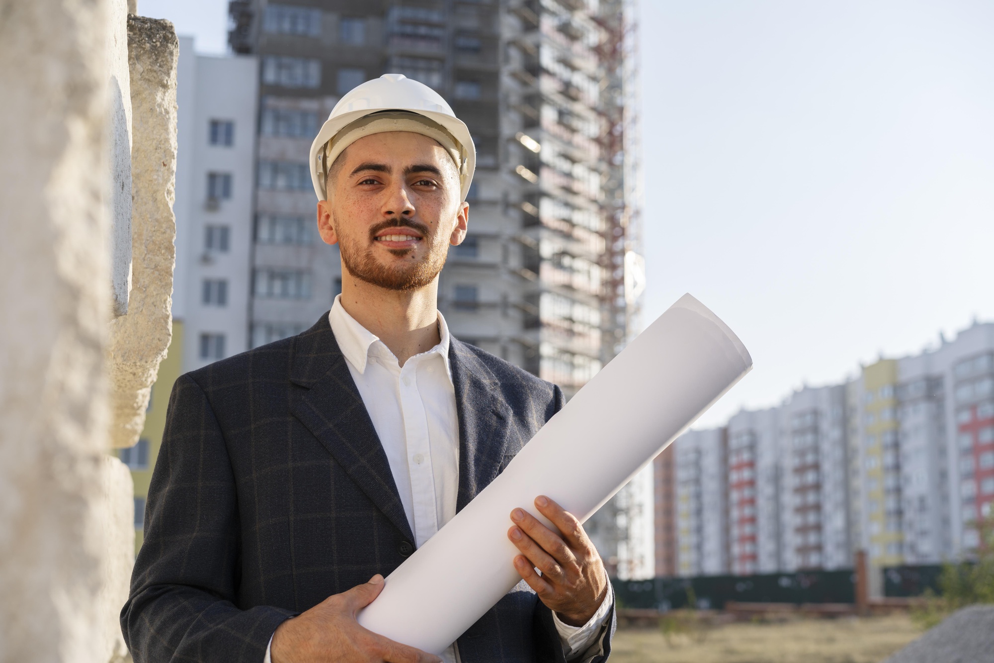 A man in suit holding a rolled floor plan wearing safety construction helmet in a construction site