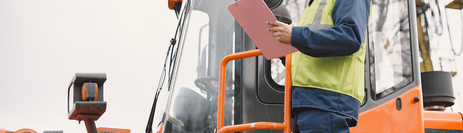 Construction site worker in safety gear inspecting heavy equipment with a clipboard
