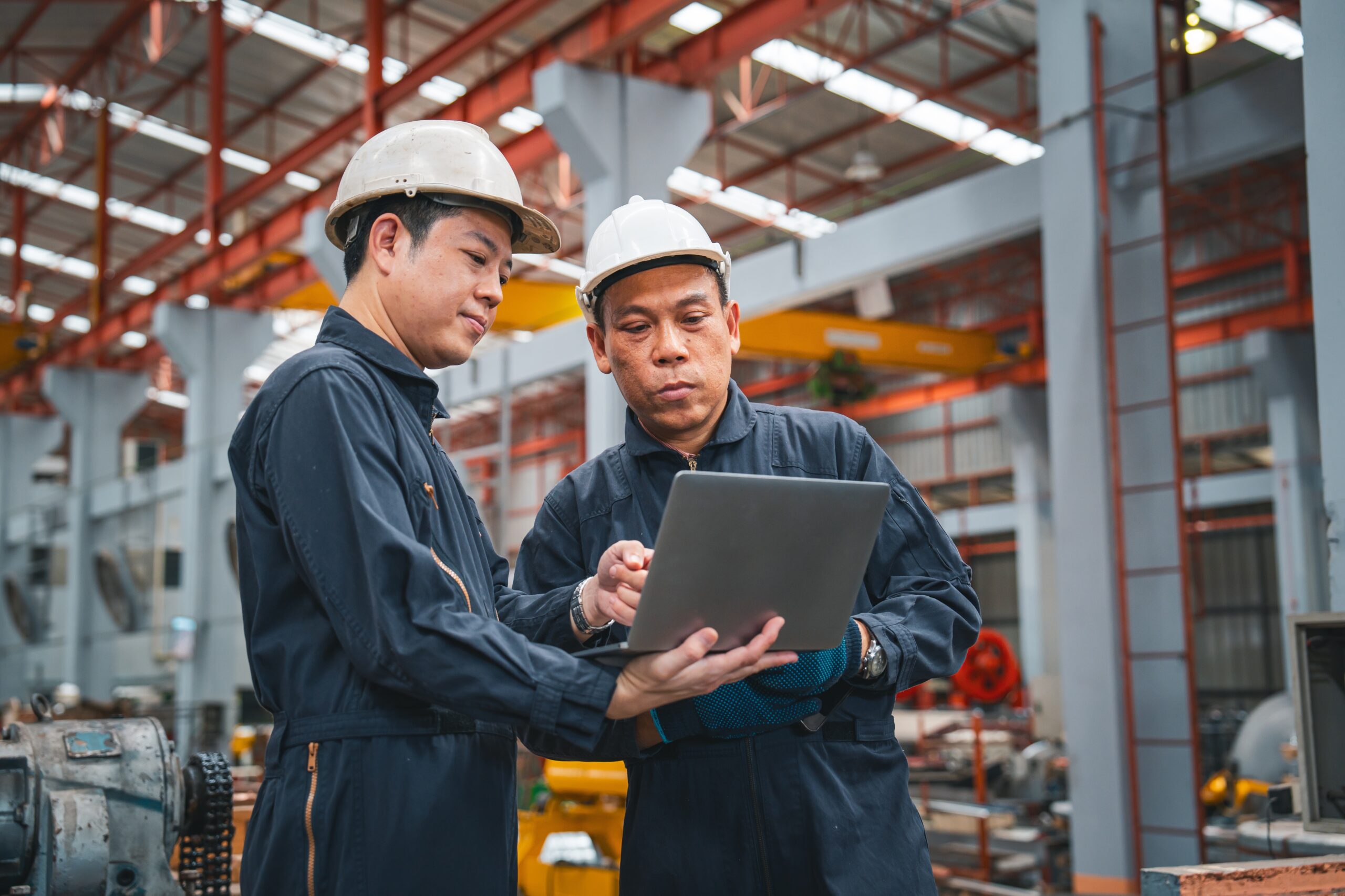 Two male engineers in white hard hats and dark navy coveralls standing in a large industrial factory or warehouse, reviewing data together on a laptop computer.