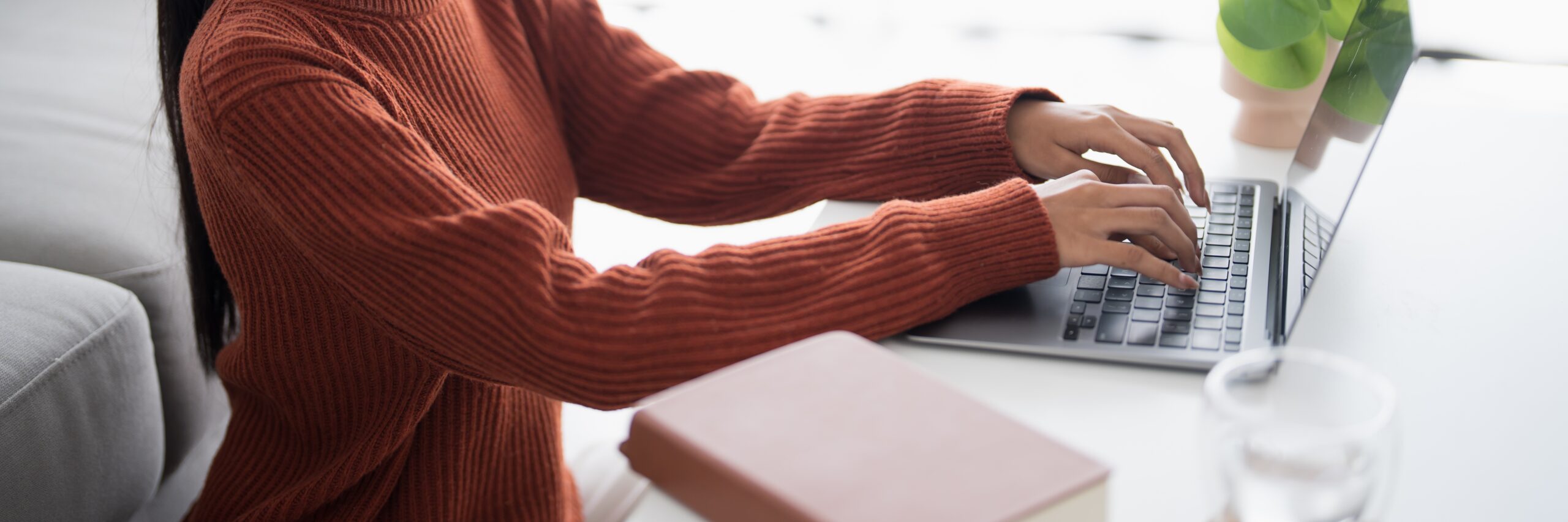 Close-up of a person's hands typing on a laptop keyboard while working from a home office or casual workspace. They are wearing a rust-colored sweater and there is a notebook and glass of water on the desk.