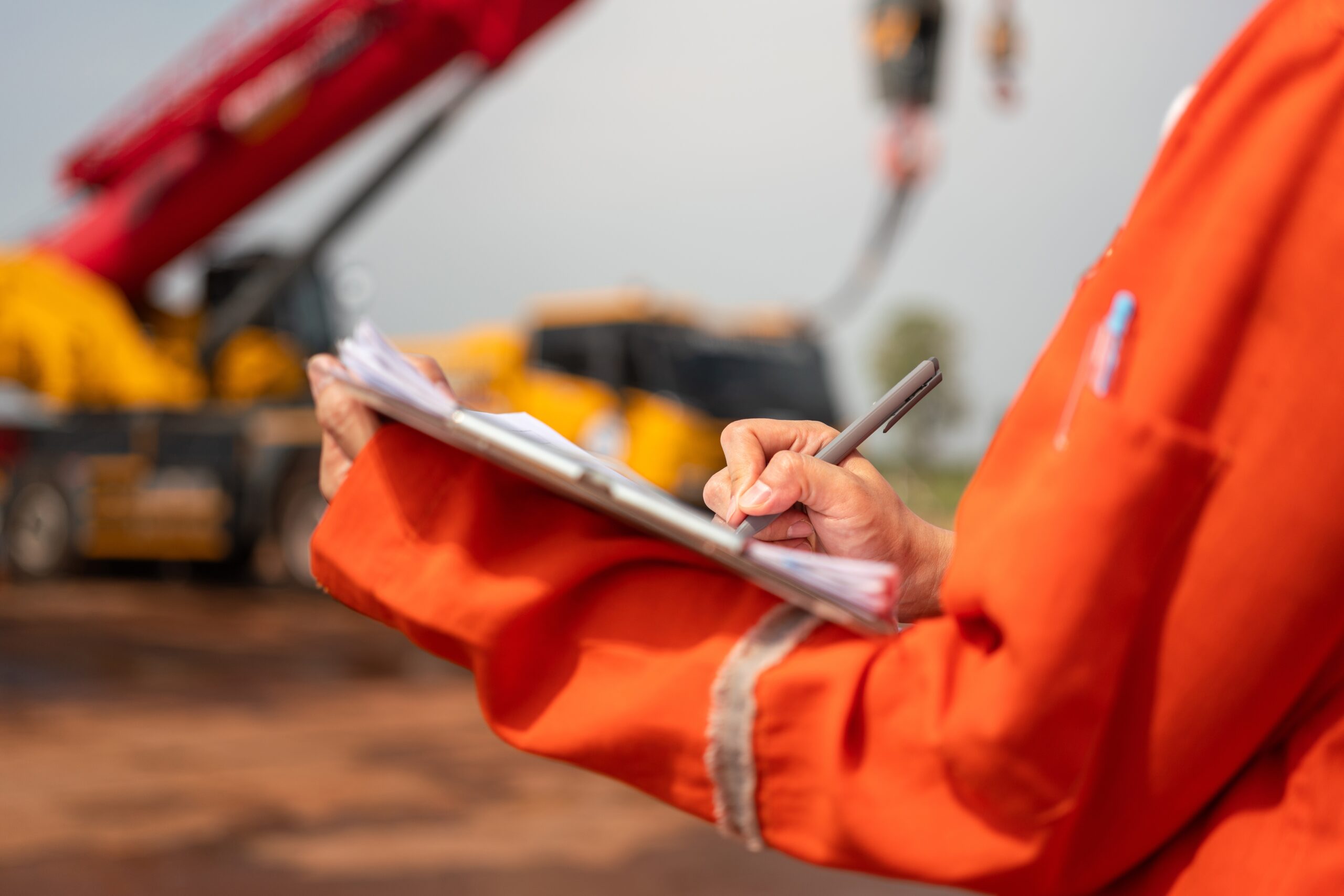 Close-up of a site supervisor in an orange high-visibility uniform writing a report on a clipboard with heavy construction machinery and a crane blurred in the background.